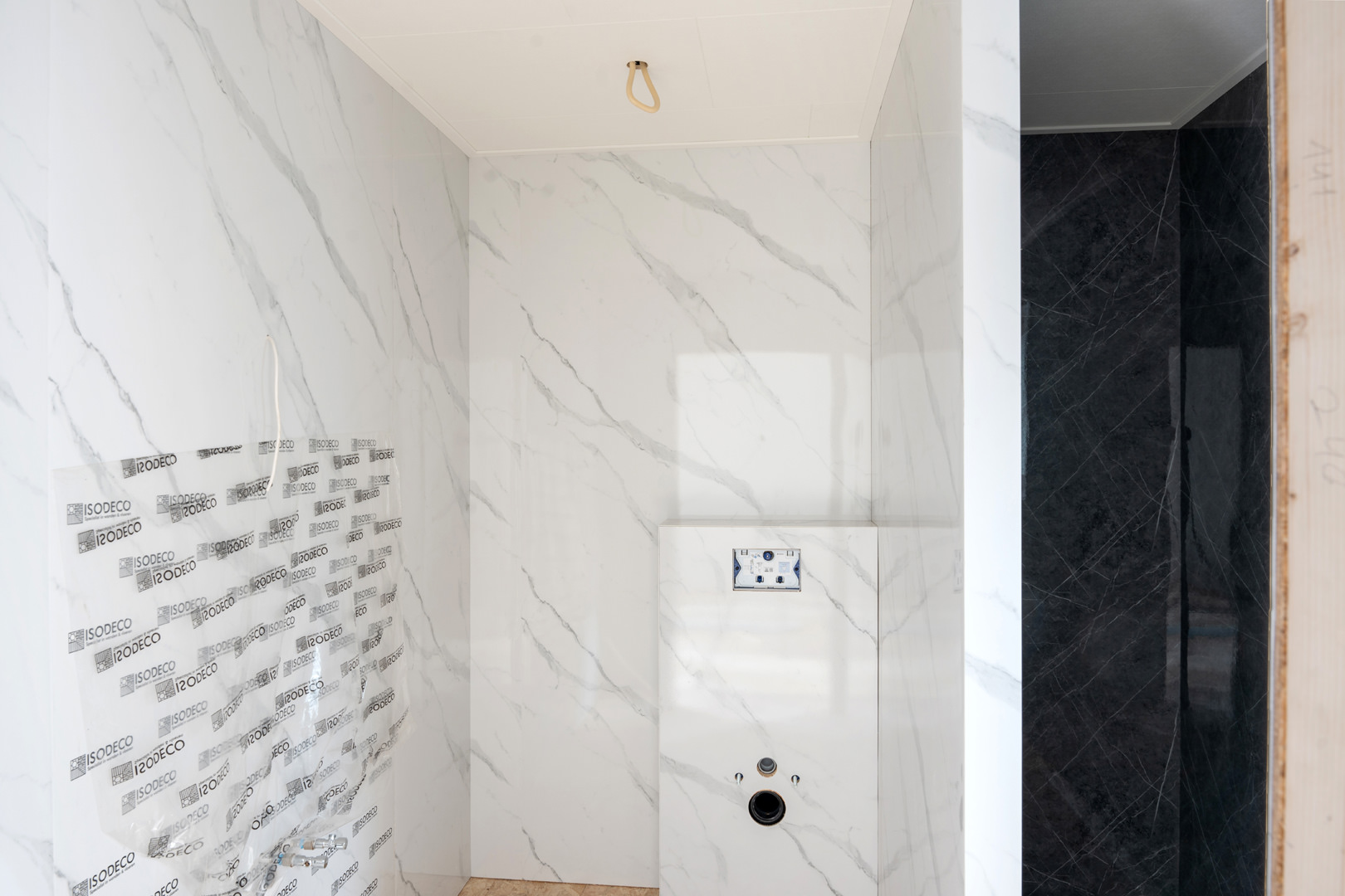 De onafgemaakte badkamer met witte marmeren muren, blootliggende bedrading en ontbrekende armaturen staat naast een donker betegelde ruimte. De scène lijkt op een ruw canvas in een fotografieproject, wachtend op de perfecte transformatie.
